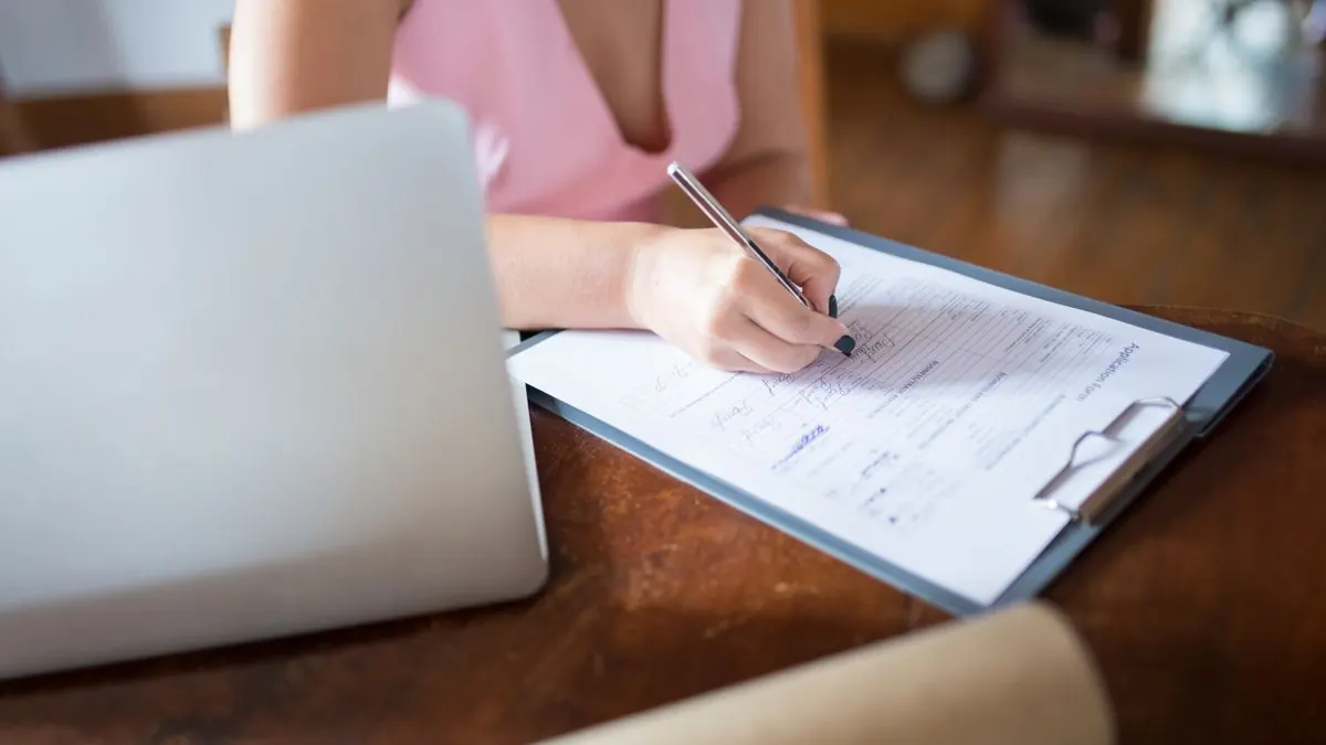 Žena vyplňuje formulář na klipboardu vedle notebooku