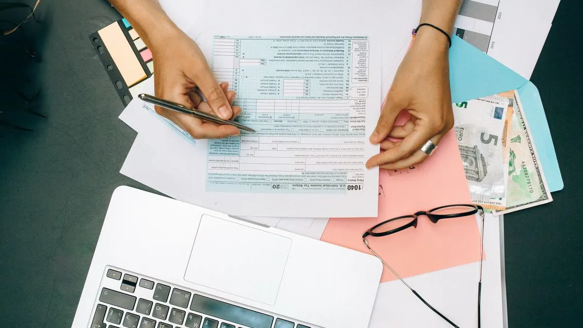Ruce vyplňují dokumenty vedle notebooku a brýlí