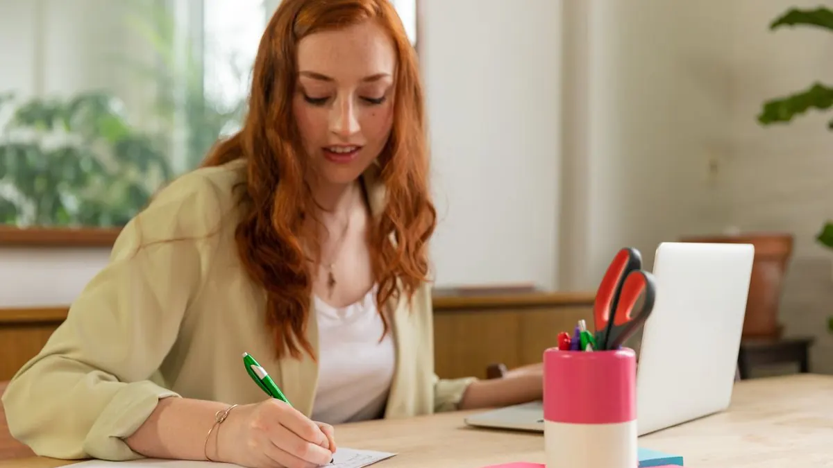 Žena píše perem na papír u stolu s notebookem