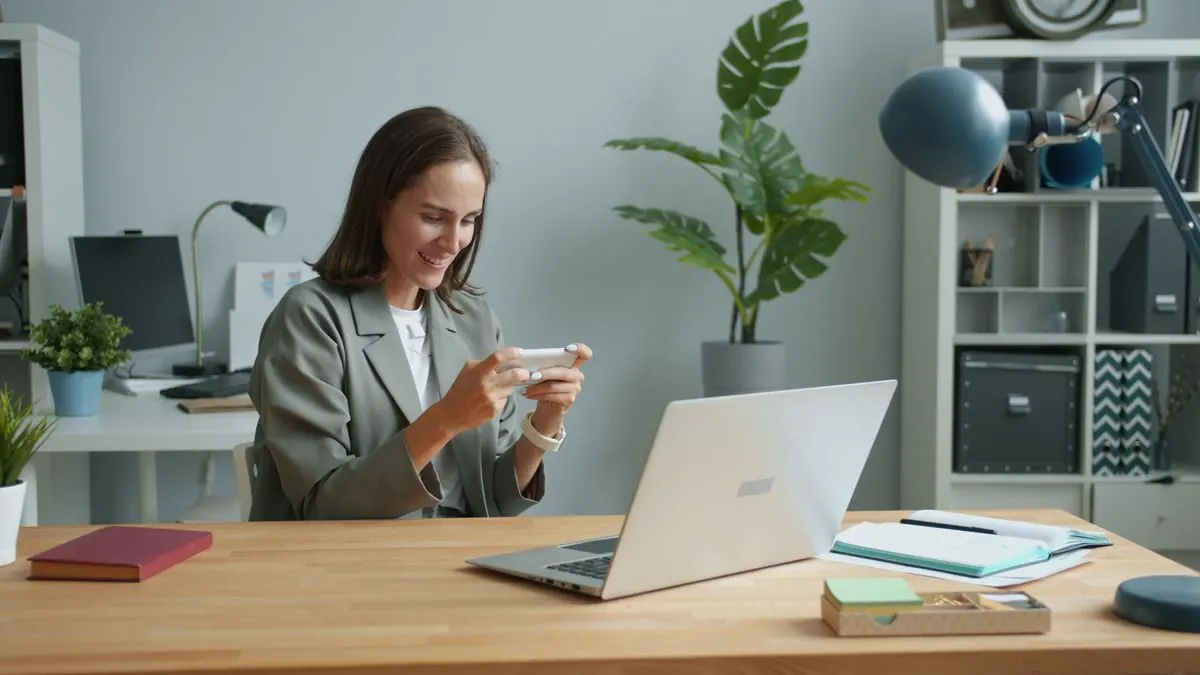 Žena v kanceláři pracuje s telefonem u notebooku, v pozadí rostlina
