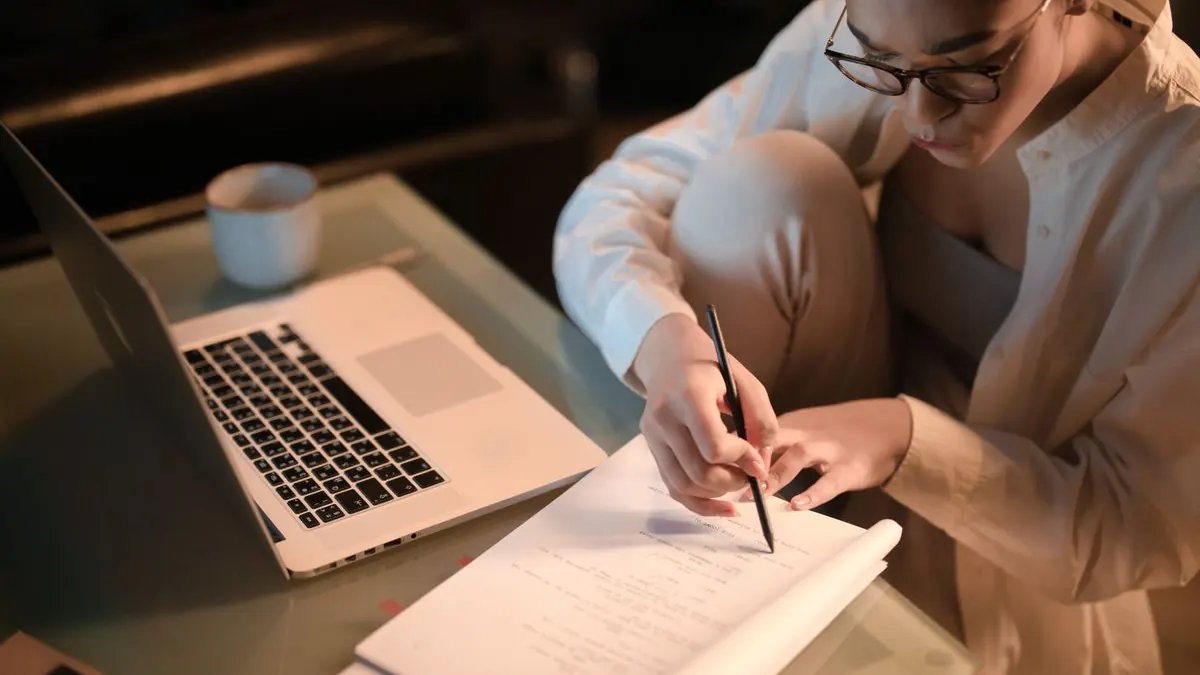 žena sedí se zkříženýma nohama a píše do sešitu vedle notebooku a kávy