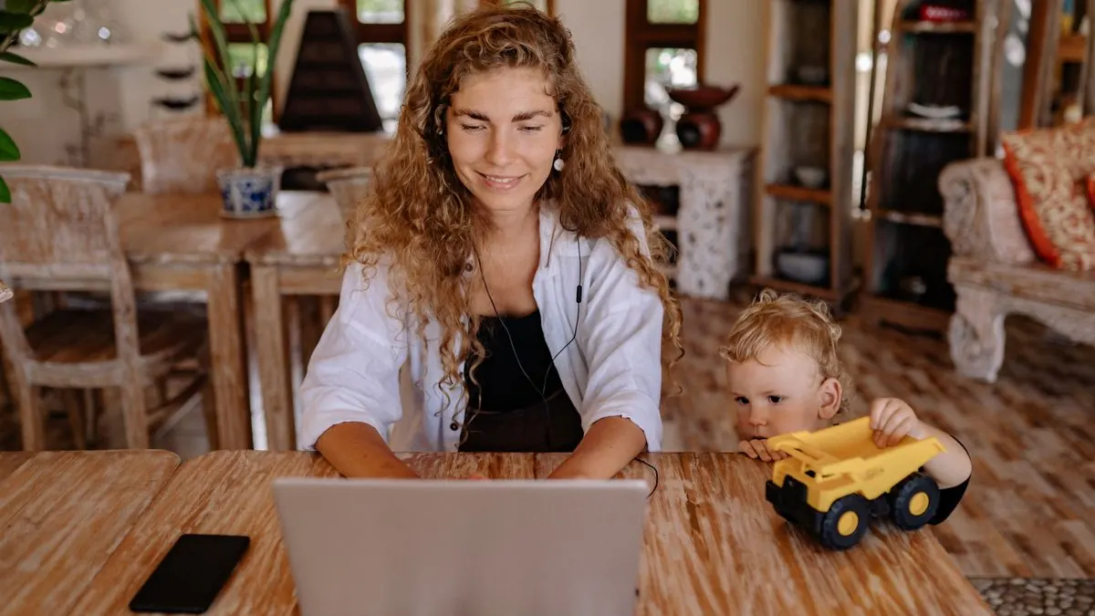 Žena pracuje na notebooku, vedle si hraje malé dítě s autíčkem