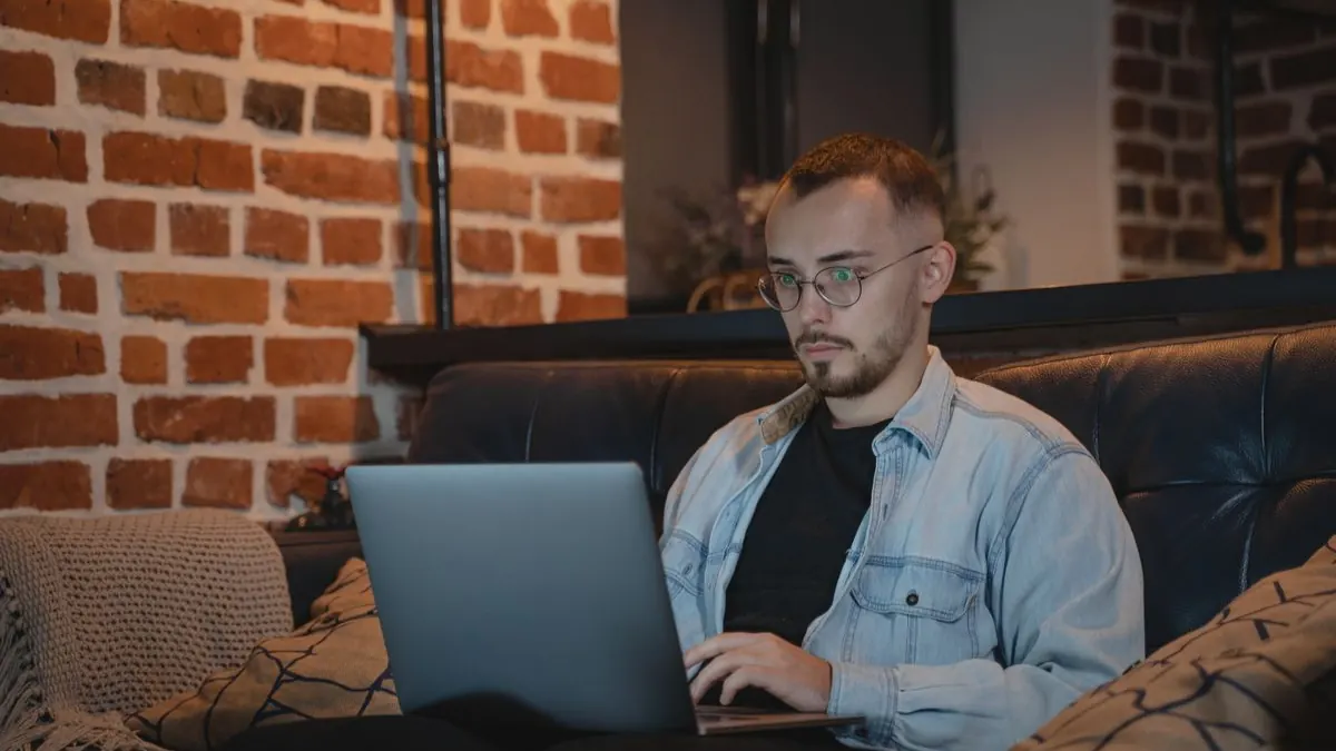 Muž sedí na gauči s notebookem u cihlové zdi