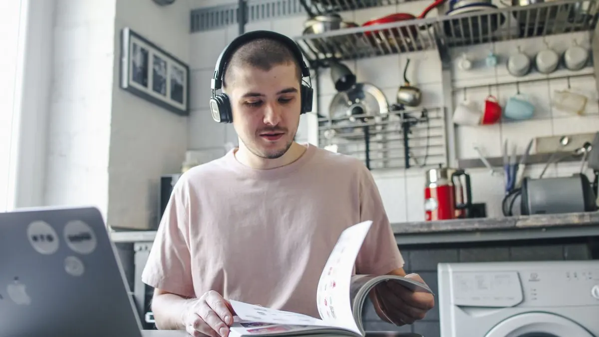 Muž se sluchátky pracuje s knihou a notebookem v kuchyni