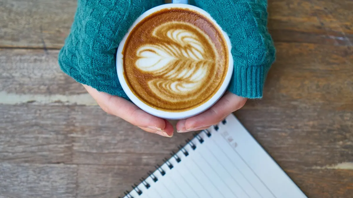 Ruce drží šálek kávy s latte art nad zápisníkem
