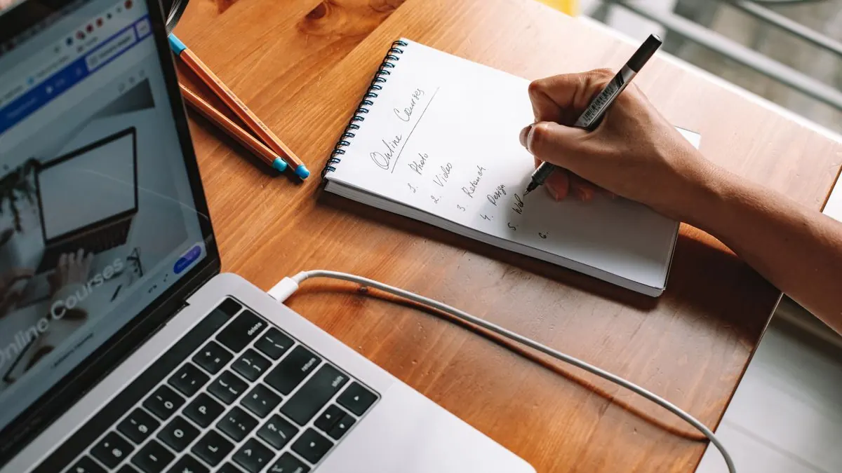 Ruka píše poznámky tužkou vedle otevřeného notebooku